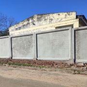 Vê-se um muro pré-fabricado e, atrás dele, o prédio da escola, pintado em amarelo e com a tinta bastante gasta. Há árvores sem folhas à esquerda. Em primeiro plano, a rua. O céu está azul, sem nuvens. 