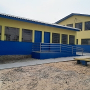 Foto externa dos prédios da escola. Em primeiro plano, uma área de areia com um banco de cimento. Ao fundo, um prédio de um piso pintado em azul e amarelo. À direita, outro prédio nas mesmas cores, mas de dois pisos.