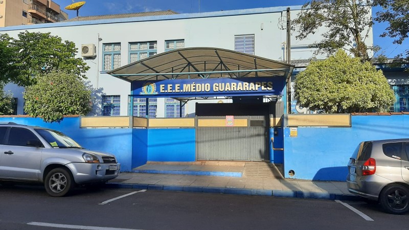 Foto externa da escola, um prédio de dois pisos em azul claro. Na rua, dois carros estão estacionados. Há um muro azul em volta da escola e uma entrada com um letreiro com o nome da escola.