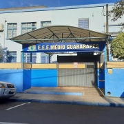 Foto externa da escola, um prédio de dois pisos em azul claro. Na rua, dois carros estão estacionados. Há um muro azul em volta da escola e uma entrada com um letreiro com o nome da escola.