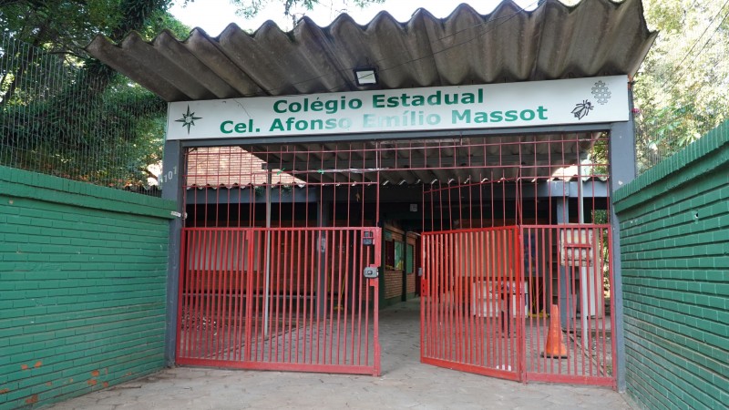 Foto da fachada da escola. Envolto por muros verdes, vê-se o portão vermelho da escola, encimado pelo letreiro com o nome da instituição.