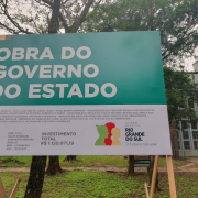 No pátio da escola, vê-se a placa indicando a obra do governo do Estado e, ao fundo, árvores e o prédio escolar.