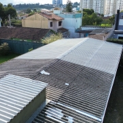 Foto aérea do prédio escolar. Vê-se a cobertura da escola e, dos lados esquerdo e direito, áreas de grama. Ao fundo, casas e prédios.