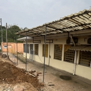 Vê-se a terra remexida em frente ao prédio de um piso da escola. Ao fundo, um poste de luz e carros.