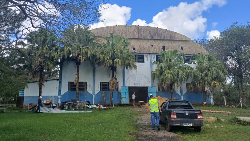 Foto externa da frente do ginásio. Há, em primeiro plano, um gramado e, no meio dele, um caminho de pedra brita. Por esse caminho, anda um homem vestido com material de proteção, tendo ao seu lado um carro. Em frente ao ginásio, há dois homens, um à esquerda e outro ao centro. Também há materiais, como cadeiras, um monte de areia e palmeiras. O ginásio é pintado em dois tons de azul. Há árvores nos lados do ginásio e o céu está azul com nuvens.