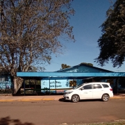 Foto externa da escola. Em primeiro plano, há a rua asfaltada e um carro branco em frente à entrada do terreno da escola. Ao fundo, a escola pintada em azul claro e duas árvores grandes.