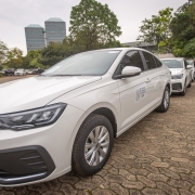 Foto de um carro sedã branco com outros carros iguais parados atrás dele.