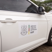 Foto em close da porta de um carro branco com um adesivo onde se lê "Serviço Público Estadual - SOP". Outro carro branco é visto em frente.