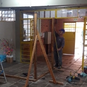 Foto interna de um corredor da escola. Um homem vestido de uniforme e capacete coloca uma escora no portão interno.