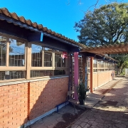 Foto da fachada da escola. O prédio é de um piso, com tijolo à vista. Ao fundo, carros e uma árvore grande. O céu está azul, sem nuvens.