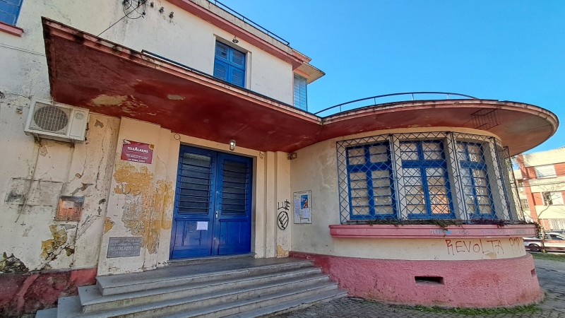 Foto da fachada da escola, pintada em bege e salmão. A escola tem dois pisos e a pintura está gasta. Ao fundo, um carro e um prédio do outro lado da rua.