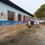 Na parte externa da escola aparecem quatro homens trabalhando, vestidos com uniforme e capacete. À direita, uma árvore. À esquerda, o prédio de um piso da escola, com parte do reboco removido, deixando os tijolos próximos ao piso visíveis. A escola é pintada em dois tons de azul. Ao fundo, um muro alto.