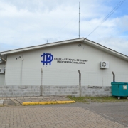 Foto externa do prédio da escola, tirada do outro lado da rua. Em primeiro plano, a rua. O prédio da escola, pintado em bege e de um piso, está depois do muro e grade. Na parede da escola, um letreiro traz o nome da instituição.