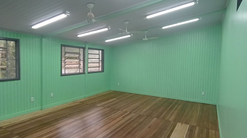 Foto interna de uma sala de aula. Ela está vazia. Suas paredes são de madeira, pintadas de verde. Há três janelas no lado esquerdo. No teto, branco, há luzes ligadas e três ventiladores.