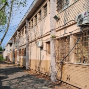 Foto externa da escola, um prédio de dois pisos pintado na cor salmão. Há janelas e ares-condicionados nas paredes. À esquerda, um caminho de pedra brita onde três carros estão estacionados. Ao fundo, árvores.