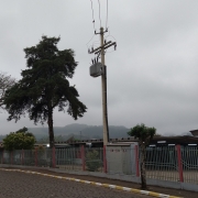 Foto da escola, tirada da rua. À esquerda, na rua de pedras, dois carros estão estacionados em frente a um ponto de ônibus. O tempo está fechado, nublado. Atrás da cerca cinza, vê-se o prédio de um piso da escola. Imediatamente atrás da cerca há três árvores e um poste de energia elétrica.