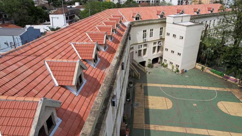 Foto aérea da altura do telhado de um prédio de dois pisos. Vê-se o telhado de telhas laranjas, o pátio da escola com árvores e uma quadra aberta de esportes. Ao fundo, os prédios da cidade.