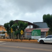 Foto da frente da escola, tirada do outro lado da rua. A escola tem dois prédio, pintados em amarelo, com uma área coberta entre eles. Há árvores na frente da escola e um carro branco estacionado. O céu está encoberto.