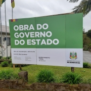 Foto da área externa da escola. Em primeiro plano, um pequeno gramado onde está instalada a placa que indica a obra. Atrás, uma área aberta e, à esquerda, o prédio da escola, de um piso e pintado em bege. O céu está encoberto.