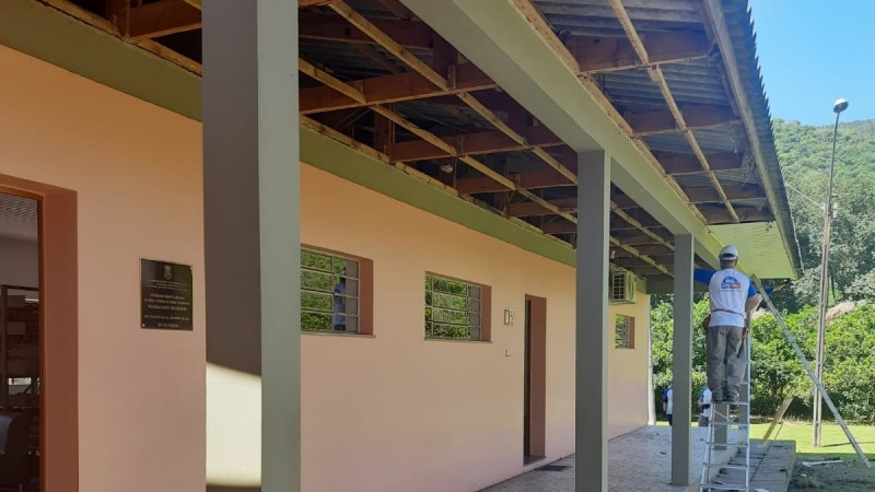 Vê-se o prédio de um piso da escola, pintada em cor salmão. Há janelas e portas no prédio, uma placa de bronze um ar-condicionado. Um homem de capacete, em uma escada, trabalha na área coberta. Ao fundo, um morro cheio de árvores.