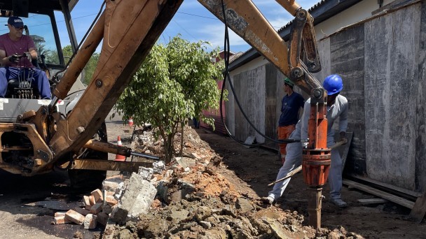 Foto externa. À direita há um prédio de um piso escondido por tapumes. Uma máquina, à esquerda, quebra o piso de cimento. Três homens trabalham. Há uma árvore pequena no centro e o céu está azul.