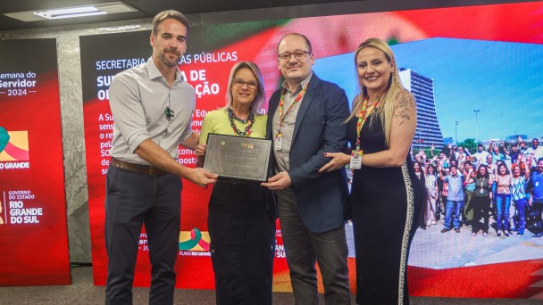 Em frente a um painel vermelho, dois homens e duas mulheres posam segurando a placa da homenagem.