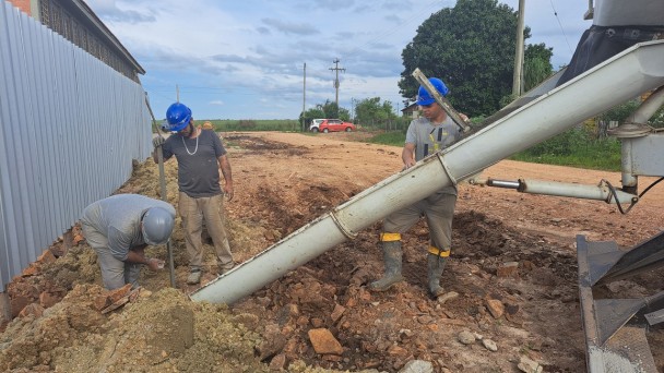 Três homens trabalham na terra. Estão usando capacetes. Há um caminhão à direita e carros ao fundo. Do lado esquerdo, um tapume separa a obra da escola.