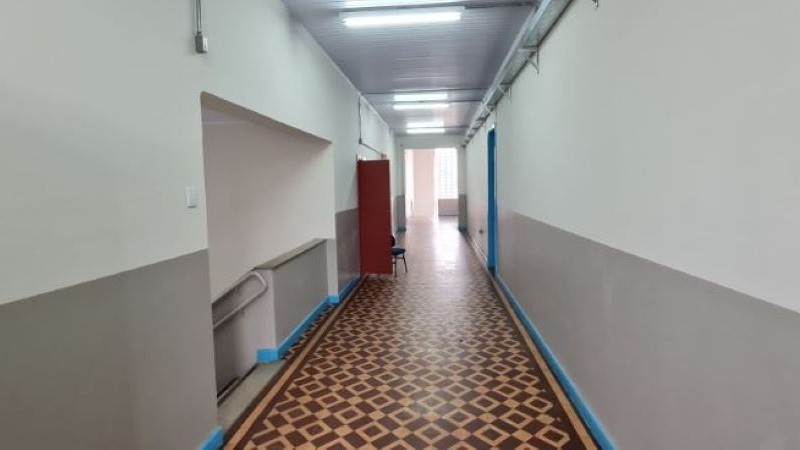 Foto interna de um corredor de escola. As paredes são pintadas de cinza e branco. Há uma porta aberta e uma cadeira no corredor. À esquerda, uma escada dá acesso ao andar inferior.