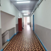 Foto interna de um corredor de escola. As paredes são pintadas de cinza e branco. Há uma porta aberta e uma cadeira no corredor. À esquerda, uma escada dá acesso ao andar inferior.