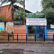 Foto externa da fachada da escola. O prédio tem dois andares em tijolo à vista. O muro é de tijolo à vista com detalhes em azul. Há um letreiro com o nome da escola em cima da entrada e, à direita, preso à tela, uma faixa. Vê-se, na frente da escola, a rua, a grade que separa a calçada da rua e um orelhão.