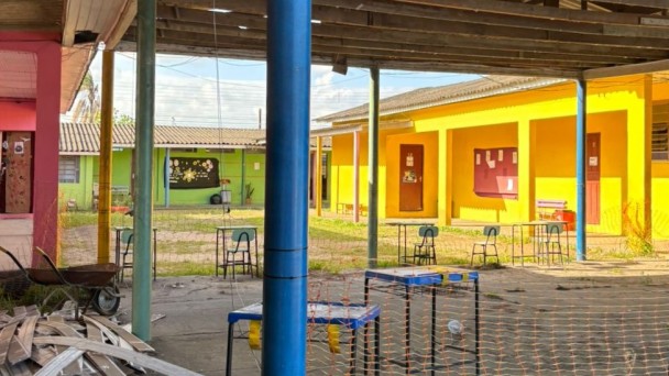 Foto do pátio interno da escola. Os prédios, em forma de U, são de um piso pintados de amarelo, à direita, verde, ao fundo, e vermelho, à esquerda. Há, em primeiro plano, uma área coberta isolada, onde há mesas e cadeiras, um carrinho de mão e materiais de construção.