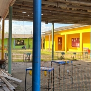 Foto do pátio interno da escola. Os prédios, em forma de U, são de um piso pintados de amarelo, à direita, verde, ao fundo, e vermelho, à esquerda. Há, em primeiro plano, uma área coberta isolada, onde há mesas e cadeiras, um carrinho de mão e materiais de construção.