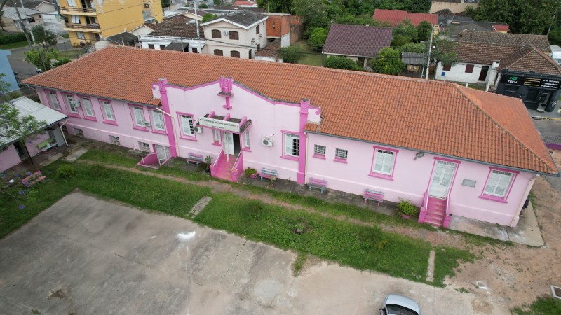 Foto aérea do prédio rosa de um piso da escola. No pátio há um carro estacionado e veem-se, atrás da escola, casas.