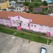 Foto aérea do prédio rosa de um piso da escola. No pátio há um carro estacionado e veem-se, atrás da escola, casas.