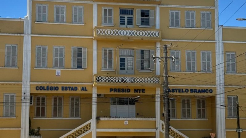 Vê-se um prédio de quatro andares, pintado em amarelo, com um letreiro onde se lê Colégio Estadual Presidente Castelo Branco, com algumas letras faltando.