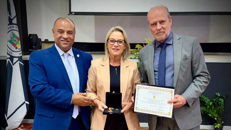 Três pessoas posam para a foto. Ao centro, uma mulher loira e, nos lados, dois homens carecas. A mulher segura uma medalha e o homem da direita um diploma. Ao fundo, se vê a bandeira da Polícia Civil.