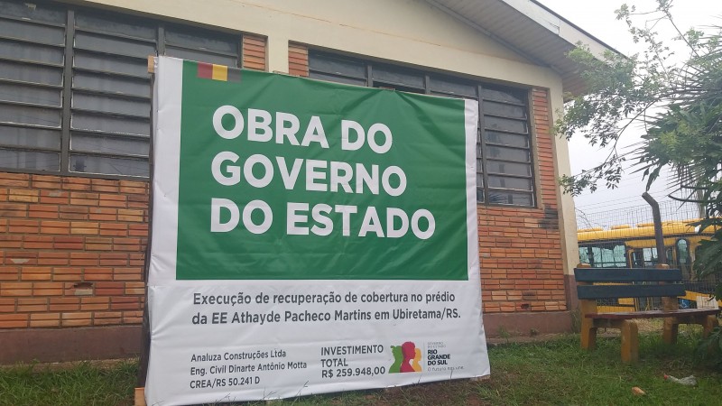 Foto de um prédio de um piso, com paredes de tijolo à vista e duas janelas. À frente, uma placa verde indica a obra do governo do Estado. Há um banco no canto direito e, fora do terreno da escola, um ônibus escolar.