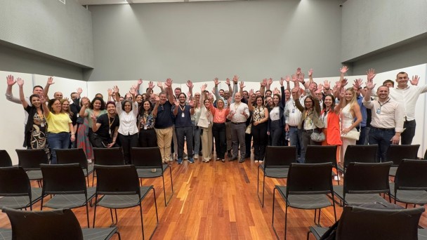 Atrás de fileiras de cadeiras, um grupo de pessoas posa com as mãos para o alto.