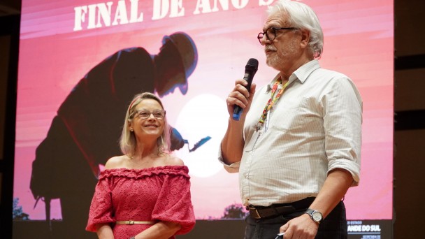 Em frente a um telão onde se vê uma foto de um homem com EPIs trabalhando com uma pá de cimento, uma mulher loira de vestido vermelho, à esquerda, olha sorrindo para um homem grisalho de cabelo branco que segura um microfone.