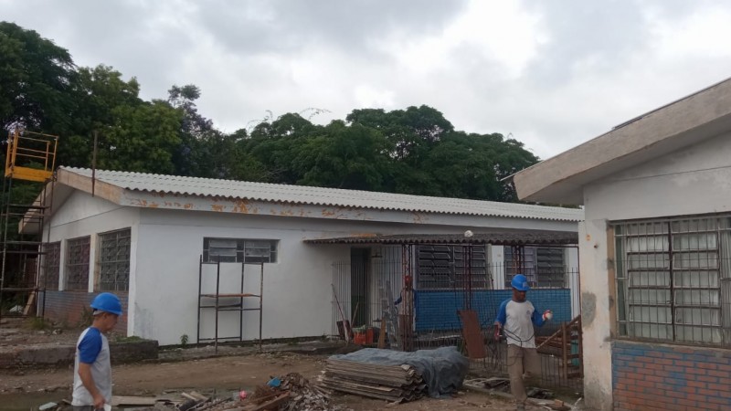 Veem-se dois prédios térreos da escola, com sinais de obra em andamento. Há três homens com capacete de obra trabalhando e um andaime à esquerda. Ao fundo, topos de árvores. O céu está nublado.