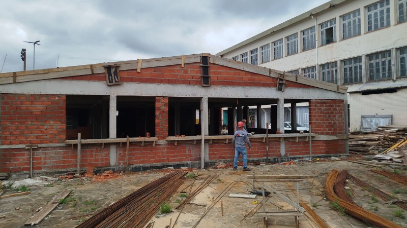 No primeiro plano, no chão, ferros de construção. Aparecem dois prédios: um de um piso, na maior parte da foto, em construção, e outro de três pisos, branco, atrás. Dois homens aparecem, um dentro da construção e outro fora.