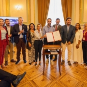 Grupo de pessoas se reúne em salão atrás de uma mesa, com o homem ao centro segurando papéis em uma pasta.