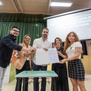 Atrás de uma mesa de escola, dois homens e três mulheres seguram uma folha de papel. Ao fundo, um homem mexe no celular.