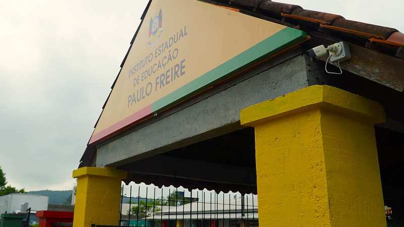 Foto da entrada da escola, com uma cobertura triangular e uma placa onde se lê Instituto Estadual de Educação Paulo Freire