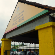 Foto da entrada da escola, com uma cobertura triangular e uma placa onde se lê Instituto Estadual de Educação Paulo Freire