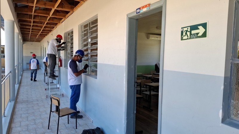 Em um corredor externo, quatro homens com capacetes pintam as esquadrias de uma escola.