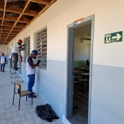 Em um corredor externo, quatro homens com capacetes pintam as esquadrias de uma escola.