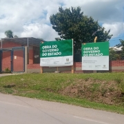 Foto externa da escola, um prédio de um piso de tijolo à vista. Em primeiro plano, se vê o asfalto da rua e uma área de grama. Há árvores na escola e placas indicando obras do estado no muro frontal. O céu está azul com nuvens.