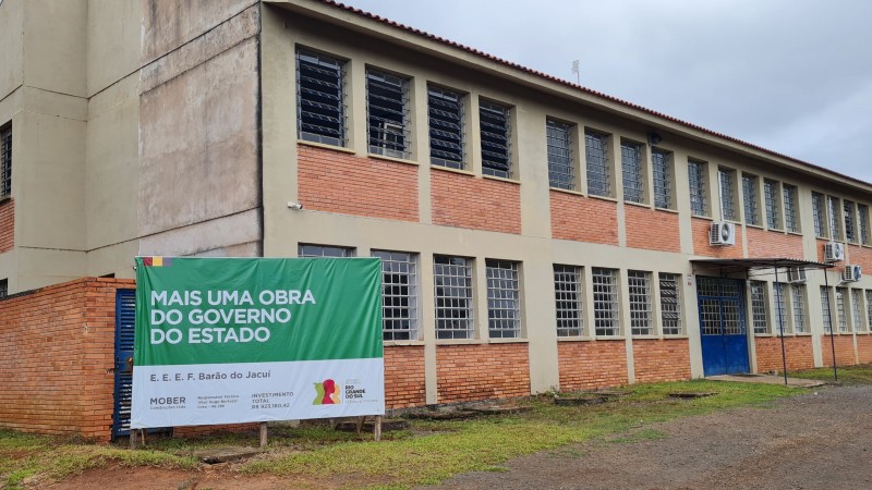 Foto de um prédio de dois pisos, com janelas nas paredes de tijolo à vista e uma placa verde indicando a obra do governo do Estado.