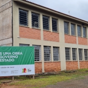 Foto de um prédio de dois pisos, com janelas nas paredes de tijolo à vista e uma placa verde indicando a obra do governo do Estado.